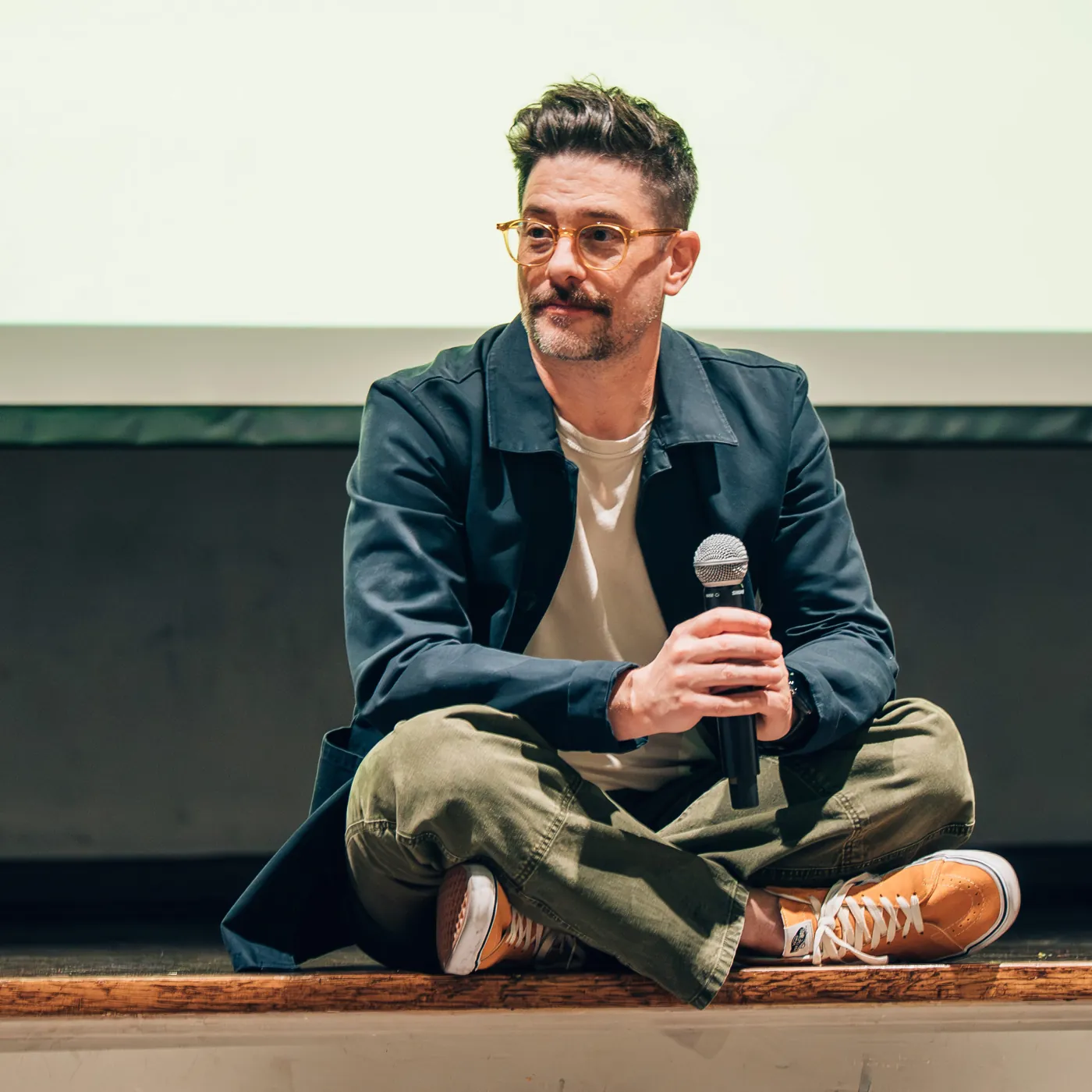 Jim Festante seated cross-legged on a stage, holding a microphone